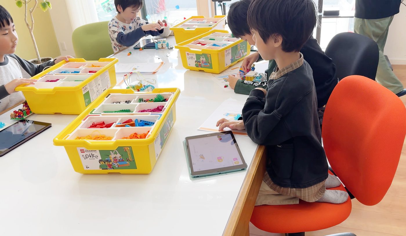 レゴ education SPIKE の教材ケースとiPadが並んだ教室の机で、レッスンに取り組む子どもたち